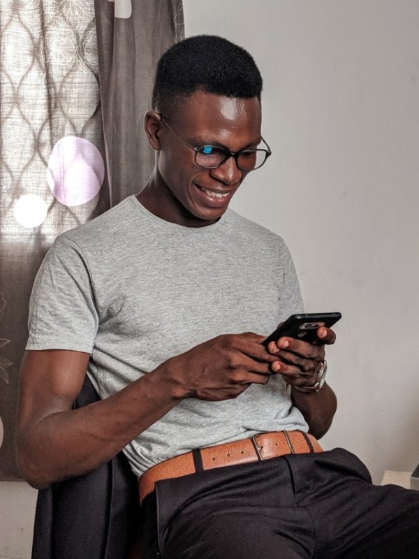 A black man wearing glasses is seated, reading something on his mobile phone.