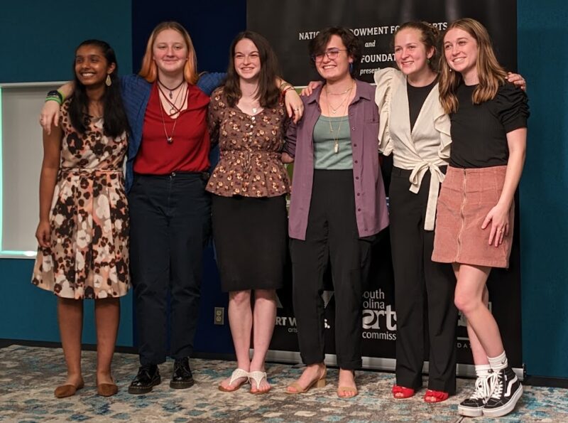 Six young women stand shoulder to shoulder with arms draped around each other on the indoor stage of the Richland Library Main Branch.