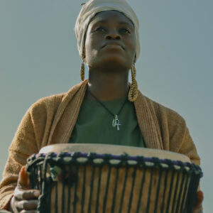 A black woman in a white head scarf holds a large handmade drum in front of her with both hands.