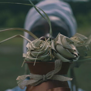 Harvested sweetgrass fashioned into a bouquet of roses in front of the out-of-focus face of a black person wearing a white head scarf.