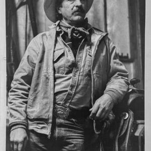 A black and white picture of a cowboy, a white male with mustache and hat standing, but leaning against a post.