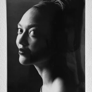 A black and white photo of a younger woman in profile with eyes cut toward the camera. Her hair is pulled back tightly.