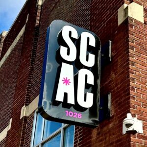 A black metal sign with white letters spelling "S-C-A-C" on the outside of our building.