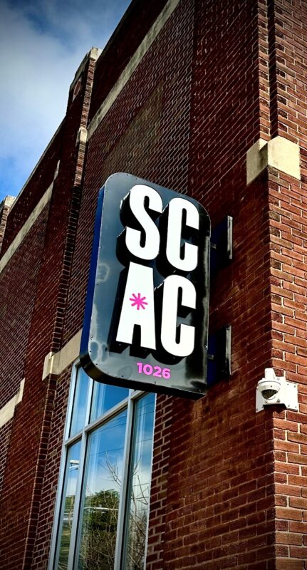 A black metal sign with white letters spelling "S-C-A-C" on the outside of our building.
