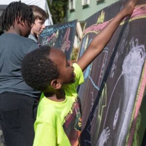 A young dark-skinned boy works on chalk artwork at an easel set outdoors at a festival.