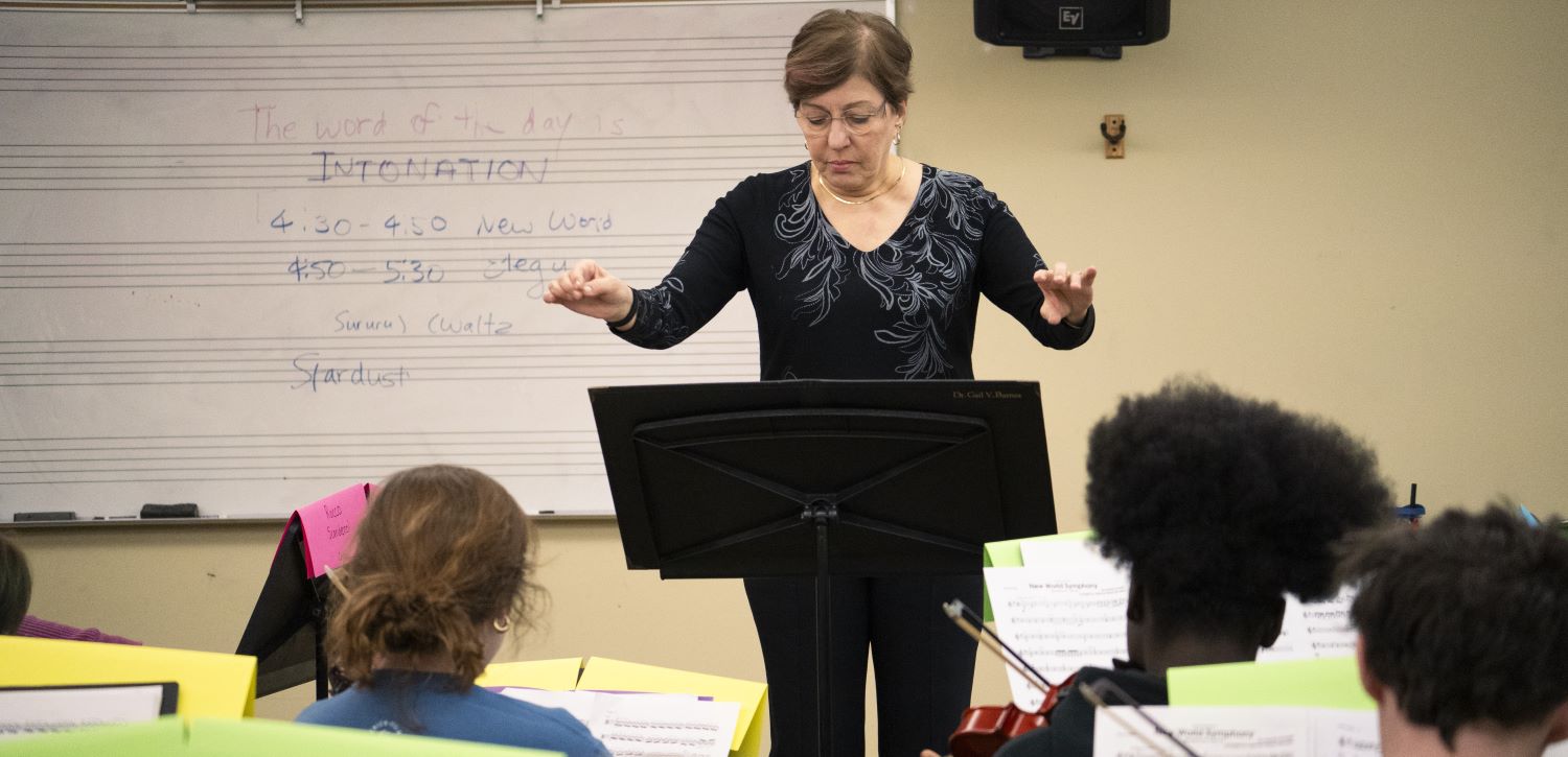 A light skinned woman with short brown hair dressed in black conducts a student string ensemble inside a studio.