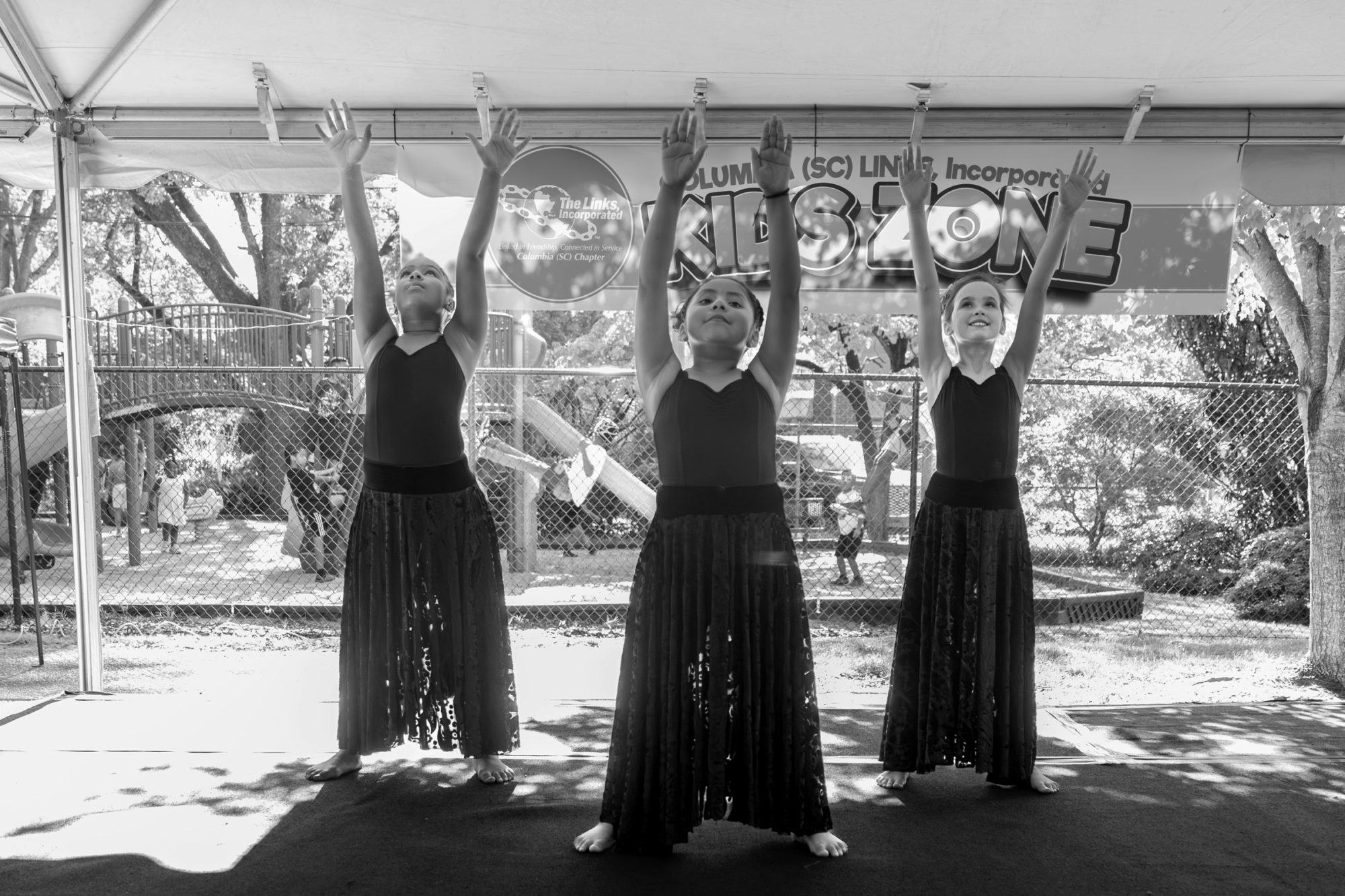 Three young female dancers pose with arms raised at an outdoor performance.