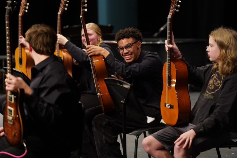 A group of male and female students on a stage in concert black attire and hold guitars they will perform.