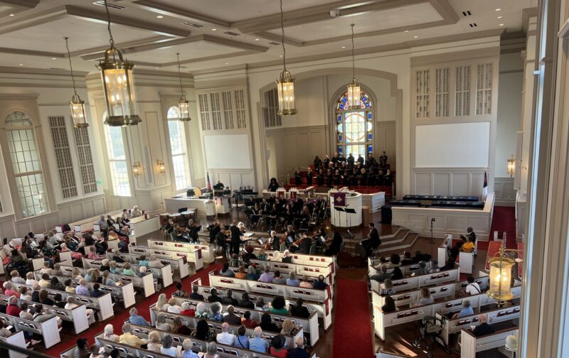 A choir and orchestra perform in a full, traditional Southern church with red carpet.