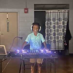A black male student stands behind a DJ turntable and sound mixing equipment.