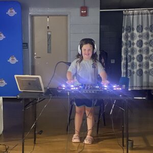 A white female student stands behind a DJ turntable and sound mixing equipment.
